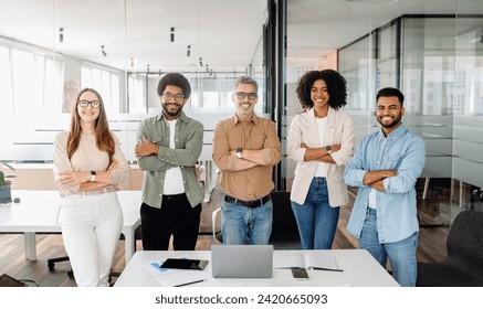 A diverse group of five colleagues stands confidently in the office, arms crossed, with smiles, symbolizing united and cooperative team ready to tackle business challenges in a co-working environment - Powered by Shutterstock