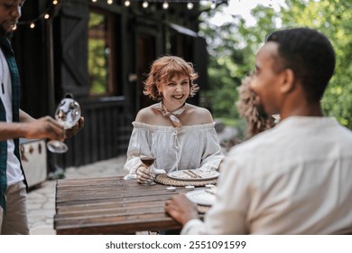 Diverse group of friends enjoying a dinner party outdoors, laughing, clinking glasses, and sharing delicious food while a waiter serves them - Powered by Shutterstock