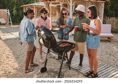 Diverse group of friends frying hot dogs on grill at beach party, drinking and talking - Powered by Shutterstock