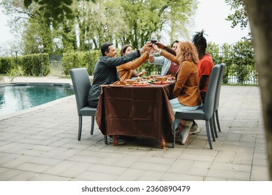 A diverse group of friends, including caucasians and an African individual, toast with wine glasses in a garden courtyard by the pool. They sit together, smiling and enjoying the moment - Powered by Shutterstock