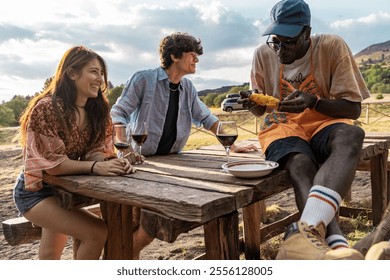 Diverse group of friends sharing wine and grilled food at rustic wooden table outdoors. Natural multicultural social gathering showing authentic connections and casual dining experience - Powered by Shutterstock