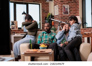 Diverse Group Of Friends Watching Scary Horror Movie On Tv, Being Afraid And Frightened, Sitting Together On Couch. People Getting Scared Of Thriller Film On Television, Drinking Beer And Bonding.