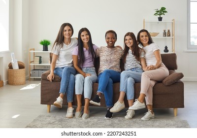 Diverse Group Of Happy Beautiful Young Women Sitting On Sofa, Smiling And Looking At Camera. Bunch Of Cheerful Mixed Race Female Friends Relaxing On Couch While Hanging Out At Home Together