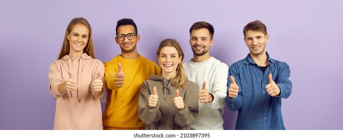 Diverse Group Of Happy Optimistic Young People Giving Thumbs Up Together. Cheerful Multiracial Multiethnic University Students Showing Thumbs Up And Smiling Standing On Purple Color Banner Background