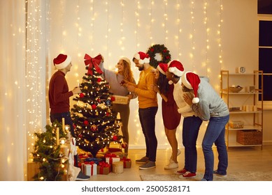 Diverse group of happy people in Santa hats having a good time at Christmas party at home. Young mixed race friends decorating their tree with beautiful, stylish baubles in a cozy, modern living room - Powered by Shutterstock