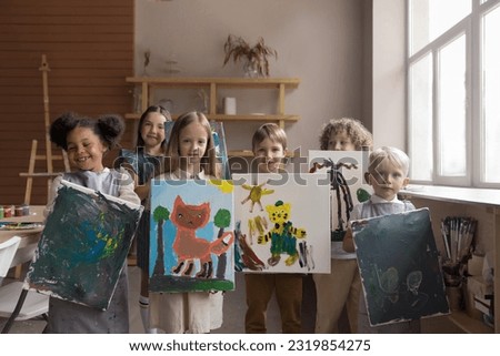 Diverse group of happy proud kids attending artistic school class, standing together, holding canvas, showing painted pictures, looking at camera, smiling, laughing, studying creativity, art