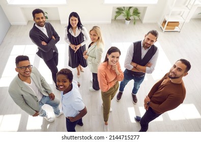 Diverse group of happy young people looking up and smiling at camera. Team of satisfied multiracial company employees standing in modern office all together. High angle indoor shot, view from above