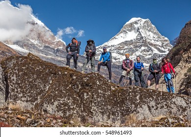 Diverse Group Of Hikers Male And Female Young And Mature Staying On Rock Holding Backpacks And Walking Sticks Mountain Valley And High Snowy Peaks On Background