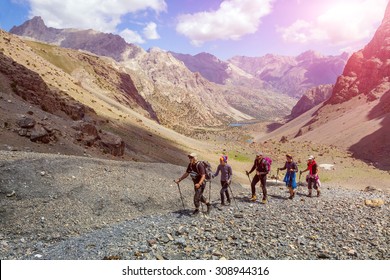 Diverse Group Of Hikers In Mountain Terrain.
People Walking Along Rocky Footpath Positive Faces Trekking Gear Poles Sporty Clothing Bright Panorama Blue Sky Sunny Day Background