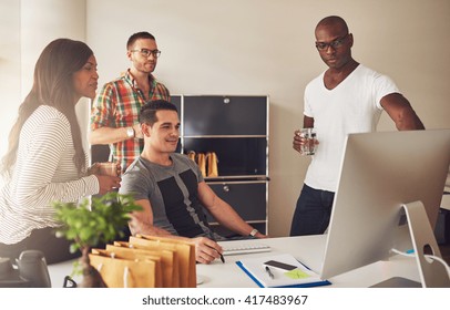 Diverse Group Of Multi-ethnic Young Adult Business People Assembled Around Desk Looking At Something On Their Computer Monitor