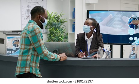 Diverse Group Of Patients With Face Masks Sitting In Hospital Reception, Talking To Receptionist About Medical Appointment. People Doing Consultation During Coronavirus Epidemic.