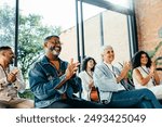 Diverse group of people attending a business meeting, happily clapping and smiling. They are seated in a bright room with large windows and natural light.