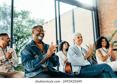 Diverse group of people attending a business meeting, happily clapping and smiling. They are seated in a bright room with large windows and natural light. - Powered by Shutterstock