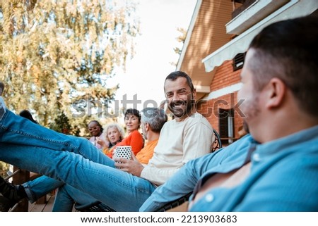 Diverse group of People hanging out, chatting and enjoying sunny autumn day in house porch. Happy Friends sitting outside in home patio, drinking mug