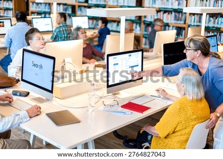 A diverse group of people in a library using computers, with a woman assisting another at a desk. Bookshelves are visible in the background.