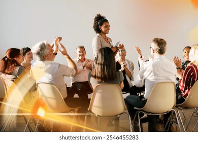 A diverse group of people meeting, including happy men and women, sitting in a circle, clapping and smiling. The group appears engaged and happy, with a woman standing and leading the session. - Powered by Shutterstock