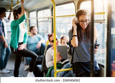 Diverse Group Of People Is Traveling By Bus With The Young Adorable Woman In The First Plan Of The Picture Who Is Smiling And Listening To Her Music.