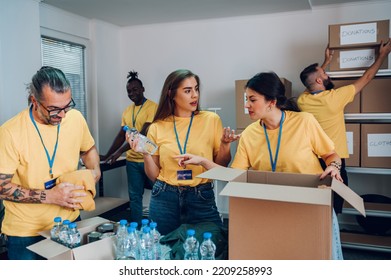 Diverse Group Of People Working In Charitable Foundation. Team Of Multiracial Volunteers In Yellow Uniform Packing Food In Cardboard Boxes And Working Together On Donation Project Indoors. Copy Space.