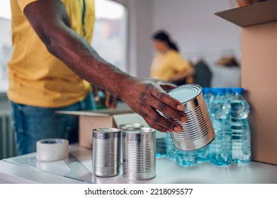 Diverse Group Of People Working In Charitable Foundation. Focus On A African American Man Hand Holding Food Can. Team Of Diverse Volunteers Working Together On Donation Project Indoors. Copy Space.