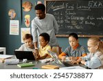 Diverse group of primary school children using laptops in pairs to study sitting at large desk in classroom while African American male teacher helping kids working on computers, copy space