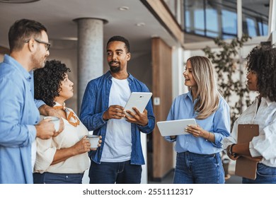 A diverse group of professionals in casual attire engaged in a lively office discussion. They are using digital tablets and coffee cups are present, indicating a collaborative work environment. - Powered by Shutterstock