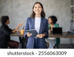 A diverse group of professionals collaborates in a modern office. One woman stands in front, confidently holding a tablet as colleagues discuss strategies and ideas, fostering teamwork and success.