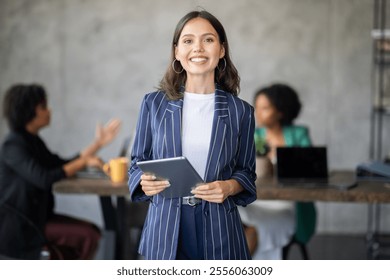A diverse group of professionals collaborates in a modern office. One woman stands in front, confidently holding a tablet as colleagues discuss strategies and ideas, fostering teamwork and success.