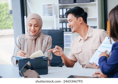 A diverse group of professionals, including a male Asian businessman and a female Muslim businesswoman in hijab, gather around a desk equipped with tablets and laptops for a productive meeting. - Powered by Shutterstock