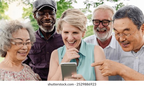 Diverse group of senior friends enjoying together and looking at smartphone. Senior men and women looking at smartphone, sharing joyful moments. Happy seniors enjoy looking at smartphone at park - Powered by Shutterstock