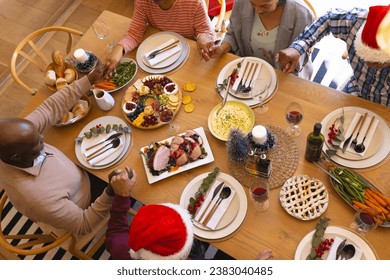 Diverse group of senior friends saying grace at christmas dinner in sunny dining room at home. Retirement, friendship, christmas, celebration, meal, senior lifestyle, communication unaltered. - Powered by Shutterstock