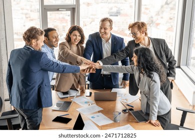 A diverse group of six business professionals stand around a conference table, their hands stacked high in the air as they celebrate a successful project. - Powered by Shutterstock