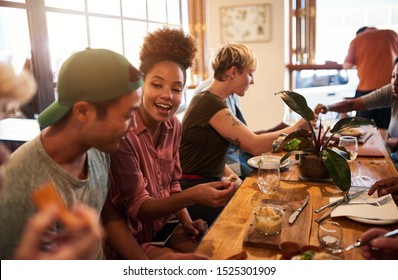 Diverse group of smiling young friends talking together over lunch and drinks at a table in a trendy bistro