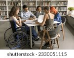 Diverse group of students inclusively college mate with disability sitting at table in library together, writing notes, working on creative essay, doing school homework together. Full length
