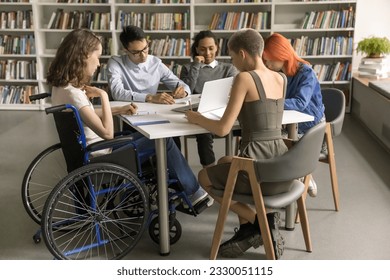 Diverse group of students inclusively college mate with disability sitting at table in library together, writing notes, working on creative essay, doing school homework together. Full length - Powered by Shutterstock