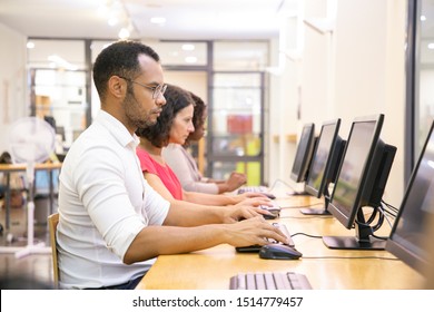 Diverse Group Of Students Taking Online Tests In Computer Class. Line Of Man And Women In Casual Sitting At Table, Using Desktops, Typing, Looking At Monitor. Training Course Concept