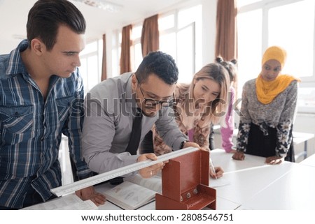 A diverse group of students at a technical college work together around a table, taking notes and measuring a red metal structural I-beam model during a civil engineering or construction class.