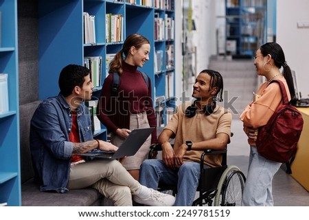 Diverse group of students with young man in wheelchair chatting cheerfully in college library, inclusivity concept