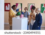 Diverse group of teenagers listening to teacher or tour guide in modern art gallery or museum