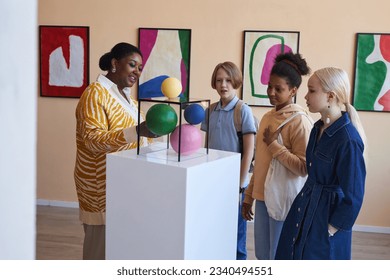 Diverse group of teenagers listening to teacher or tour guide in modern art gallery or museum