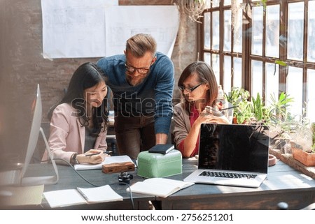 A diverse group of three professionals collaborating in a bright office, with laptops and documents on the table, surrounded by natural light and greenery.