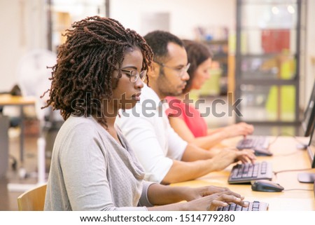 Diverse group of trainees studying in computer class. Line of man and women in casual sitting at table, using desktops, typing, looking at monitor. Computer class concept