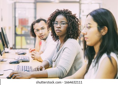 Diverse group of trainees working in computer class. Line of man and women in casual sitting at table, using desktops, looking at monitor or camera. Training concept