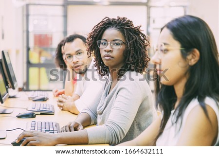 Diverse group of trainees working in computer class. Line of man and women in casual sitting at table, using desktops, looking at monitor or camera. Training concept