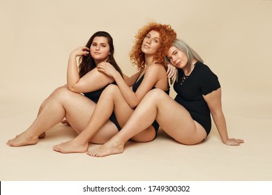 Diverse. Group Of Women With Different Figure And Size Portrait. Diversity Female Posing On Beige Background. Blonde, Brunette And Redhead In Black Bodysuits Sitting On Floor. 