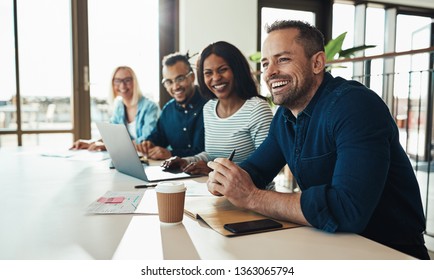 Diverse Group Of Young Businesspeople Laughing While Sitting Together In A Row At An Office Desk During A Meeting