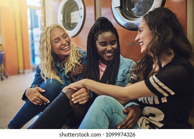 Diverse Group Of Young Female Friends Sitting On A Laundromat Floor Talking And Laughing Together While Doing Laundry