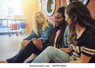 Diverse Group Of Young Female Friends Sitting On A Laundromat Floor Together Laughing And Blowing Bubbles With Chewing Gum