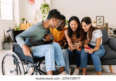 Diverse group of young friends laughing using mobile phone together at home. Inclusion concept with young indian man in wheelchair having fun with friends watching social media content on cell - Powered by Shutterstock