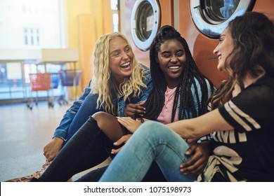 Diverse Group Of Young Girlfriends Sitting Together On A Laundromat Floor Laughing And Talking While Doing Laundry