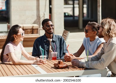 Diverse Group Of Young People Enjoying Cocktails While Sitting At Cafe Table Outdoors Lit By Sunlight